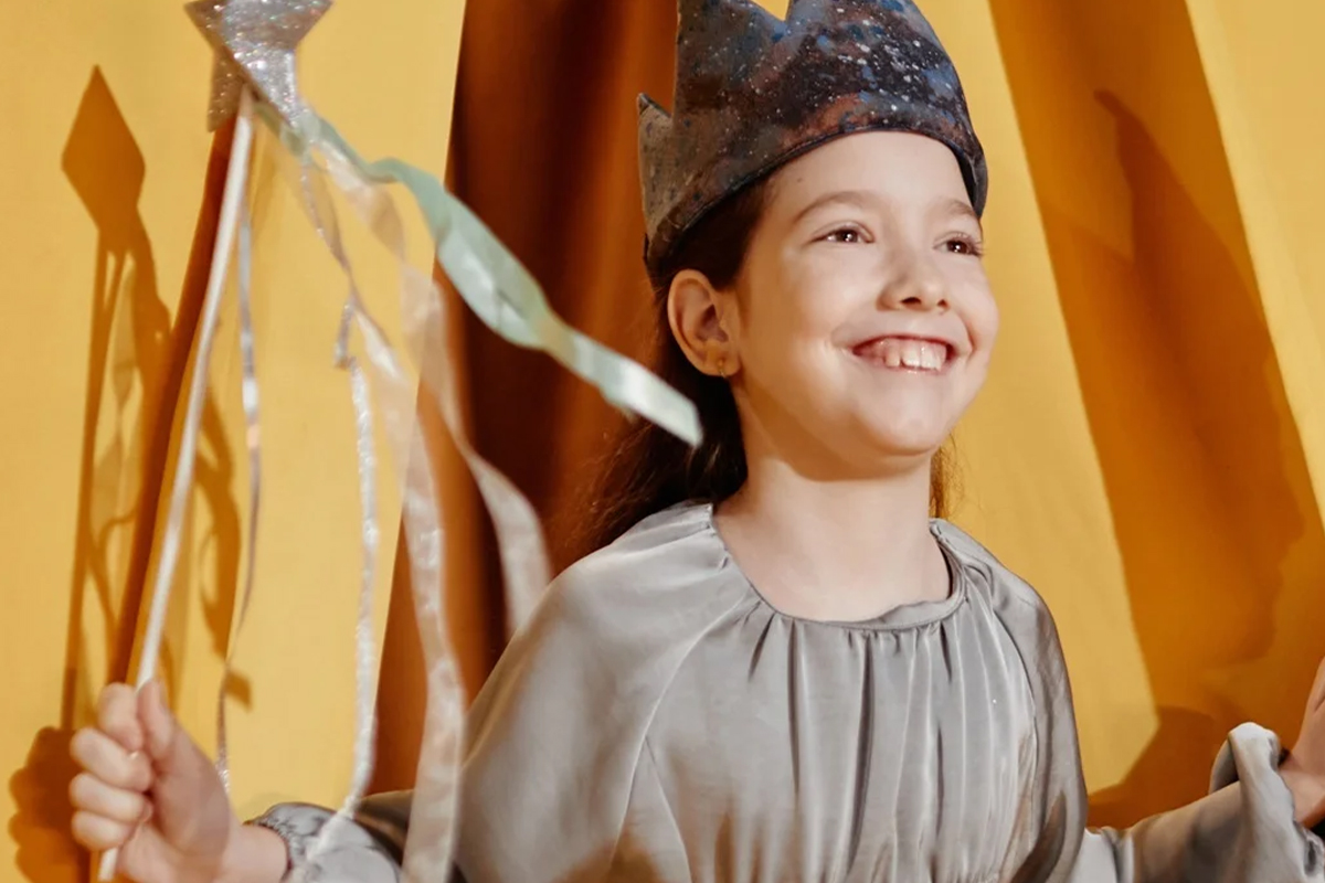 A young girl in a gray costume and handmade paper crown grins while holding a glittery wand with ribbons, performing in a children's theater production.