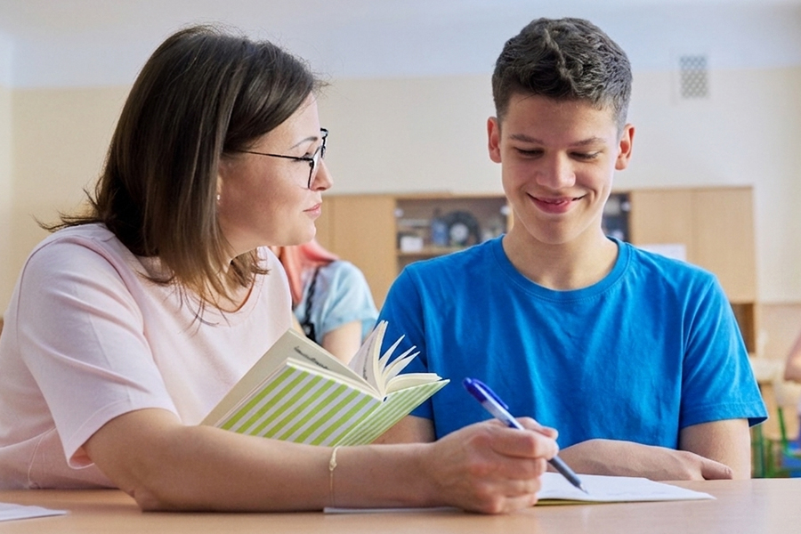 An instructor holding an open book sits beside a teenage boy who smiles while writing in a notebook. Other students are visible in the background of the classroom.