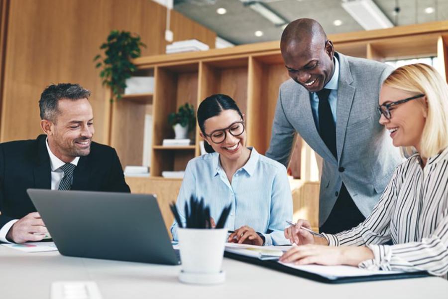 A diverse team of four office professionals in business attire smiling and collaborating around a laptop and paperwork in a modern, sunlit workspace with wooden shelving in the background.