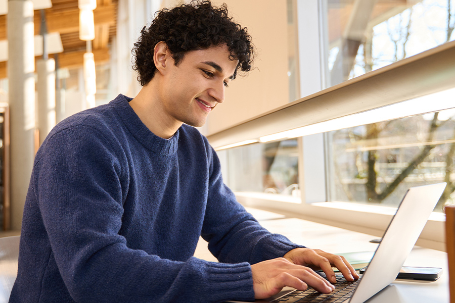 A young man in a blue sweater smiles while working on a laptop at a well-lit study space.