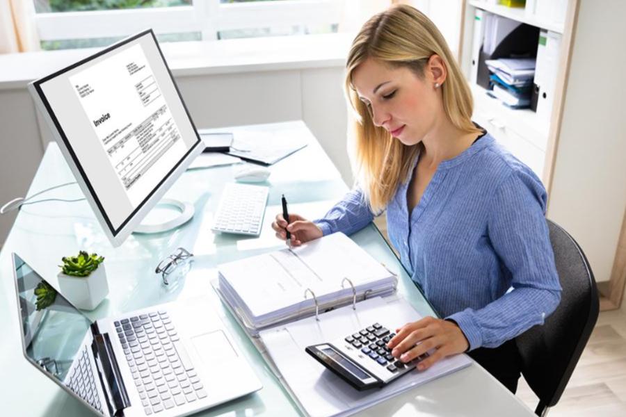 A professional woman in a light blue shirt sits at a bright, modern desk, working with an accounting calculator and a large binder of documents. Her workspace includes a laptop and a desktop monitor displaying a digital invoice, representing a focused professional environment for accounting or financial tasks.