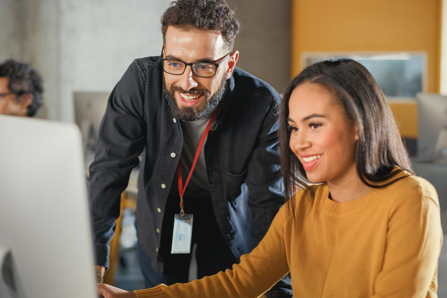 An instructor wearing glasses and a lanyard leans in to assist a smiling student at a computer.
