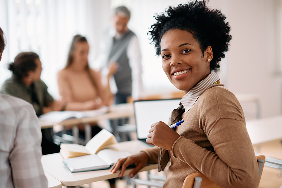 A smiling woman holding a pen looks toward the camera while seated in a classroom, with other students and an instructor visible in the background.