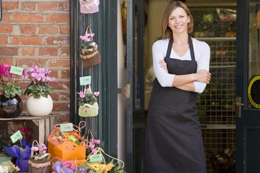 A smiling female florist in a black apron stands with her arms crossed at the entrance of a charming flower shop surrounded by potted plants.