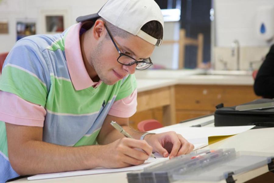 A male student wearing a white baseball cap and glasses sits at a desk, focused on drawing or sketching in a brightly lit classroom.