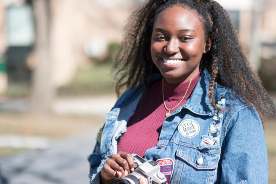 A smiling young woman in a denim jacket with patches holds a camera while standing outdoors.