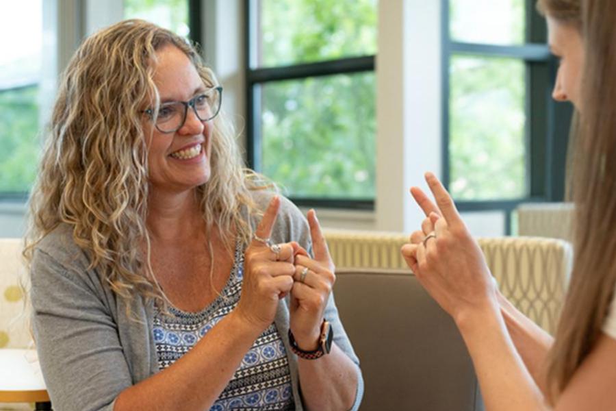 Two women smiling and communicating using American Sign Language in a brightly lit indoor lounge.