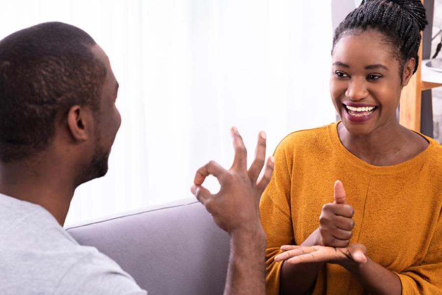 A smiling woman in a mustard yellow sweater and a man engage in a conversation using sign language.