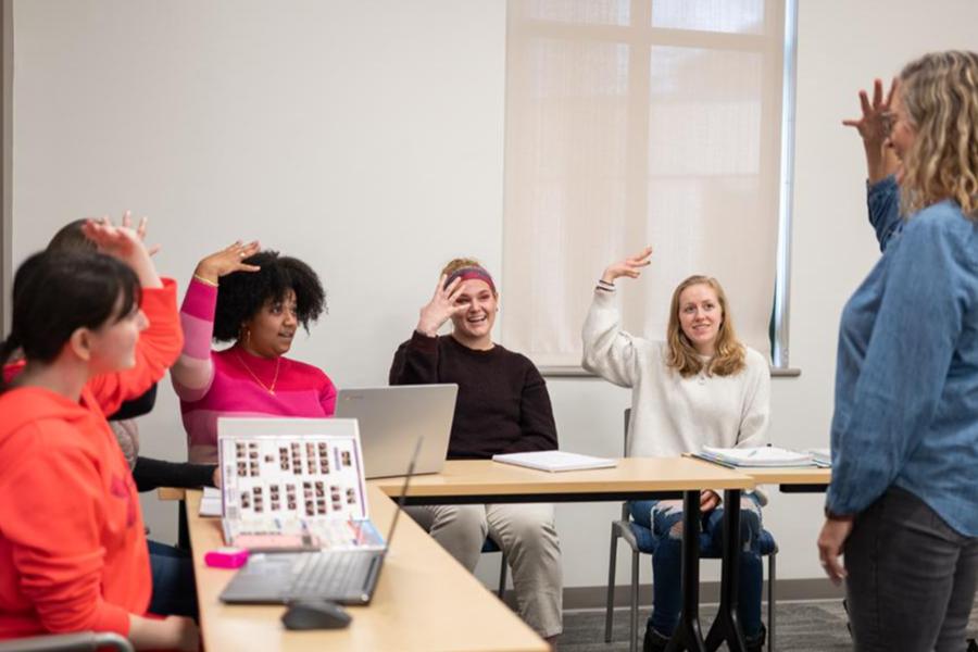 A group of students in a classroom setting learn American Sign Language (ASL) from an instructor, practicing hand signs together at their desks.