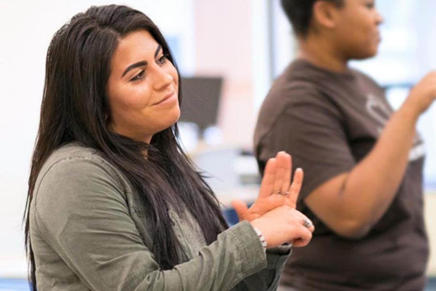 A young woman with long dark hair smiles while practicing American Sign Language (ASL) during a class or workshop.