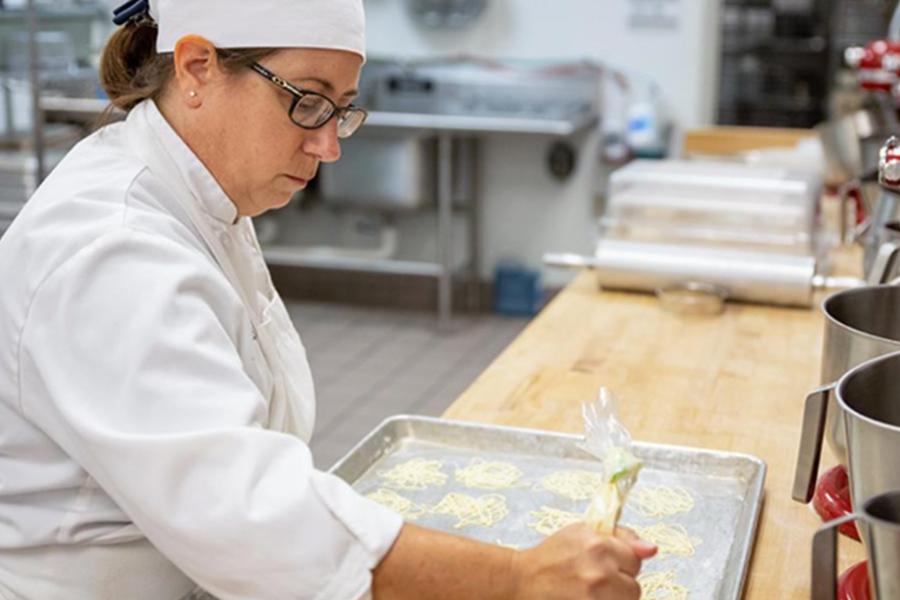 A professional pastry chef in a white uniform and hat carefully pipes delicate designs onto a baking sheet in a commercial kitchen.