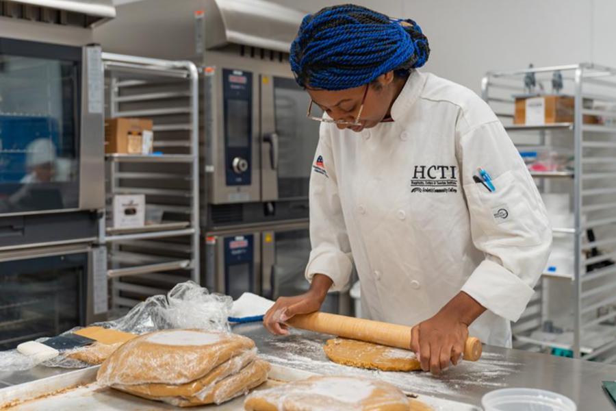 A culinary student with blue braided hair and a chef's coat uses a rolling pin to flatten dough in a professional kitchen.