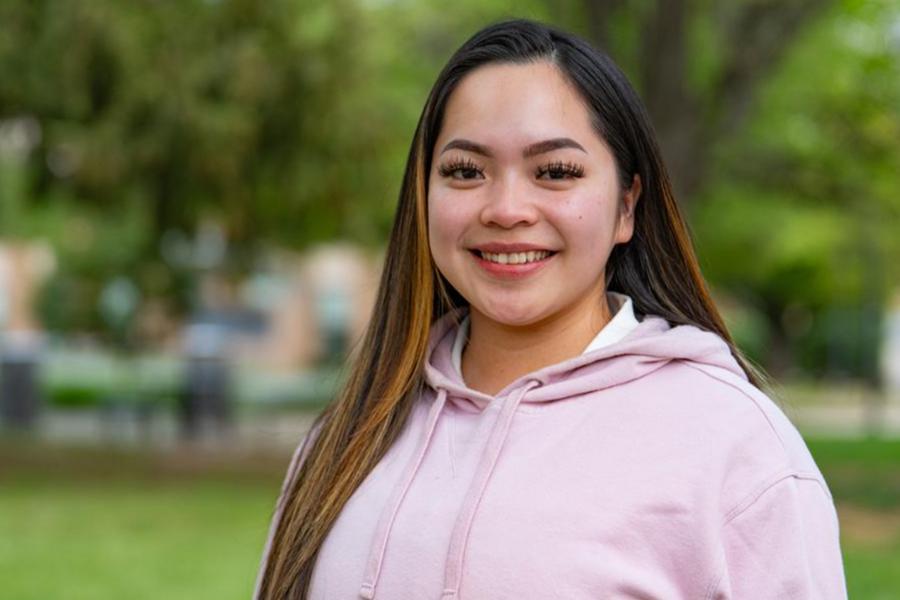 A smiling young woman with long dark hair wearing a light pink hooded sweatshirt, standing outdoors on a grassy campus with blurred green trees and college buildings in the background.