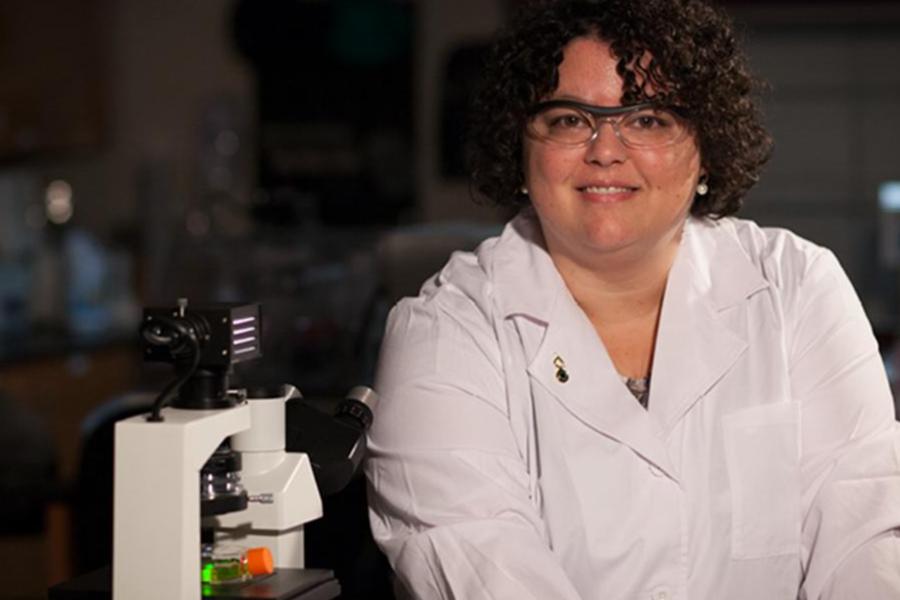 A professional portrait of a female scientist with curly dark hair wearing a white lab coat and safety glasses, smiling next to a high-powered microscope in a research laboratory.
