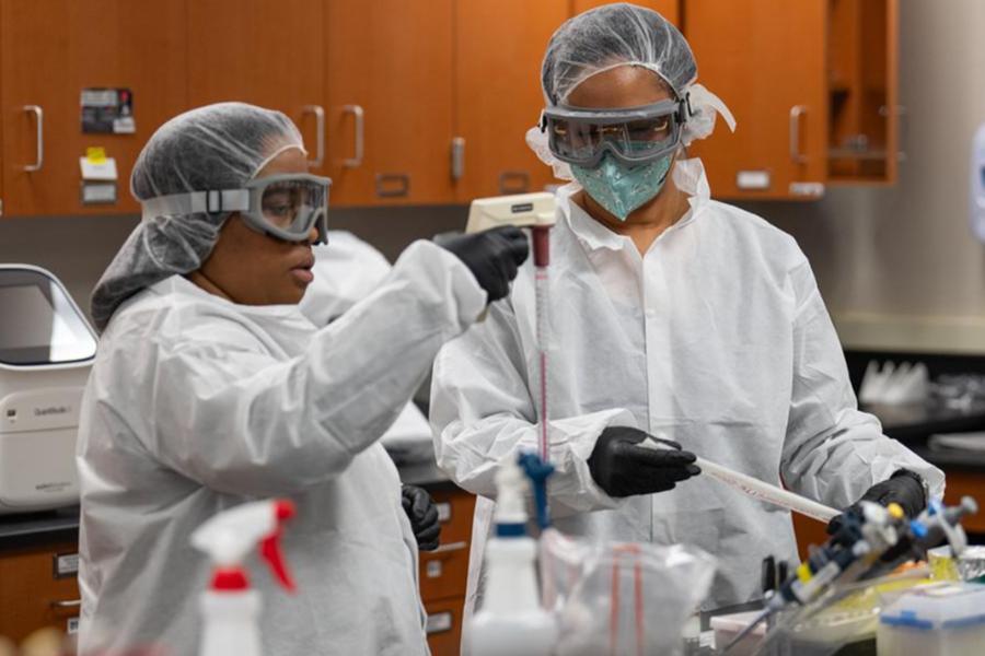 Two scientists in protective gear work in a lab, handling pipettes and samples.