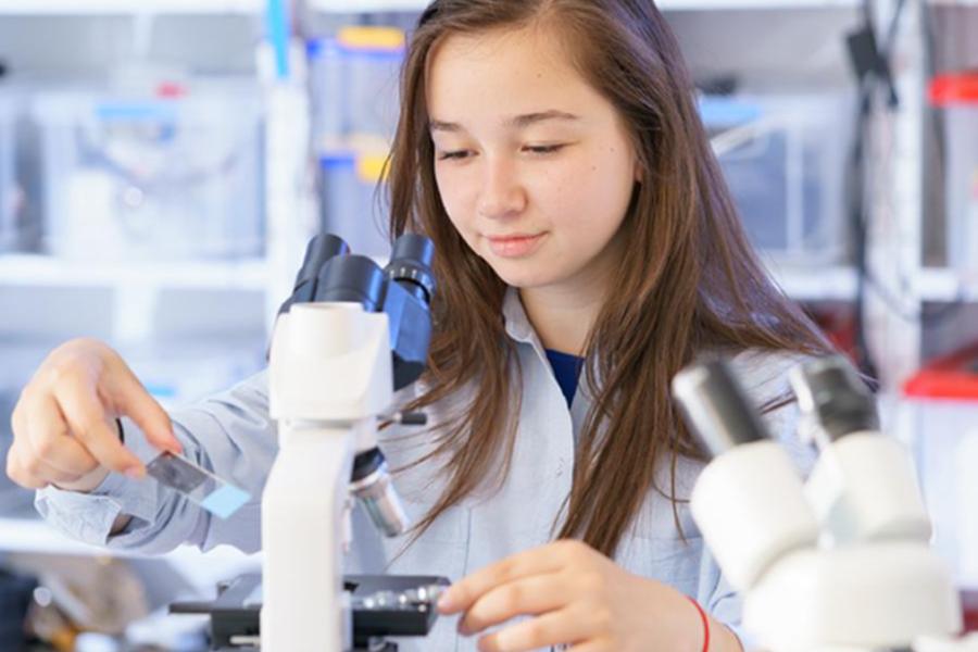 A young female student in a science lab holding a glass slide next to a white binocular microscope.