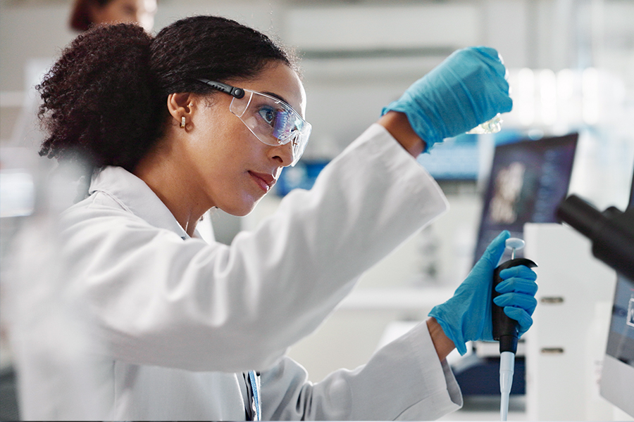 A scientist in a white lab coat, safety glasses, and blue gloves uses a pipette in a laboratory setting.