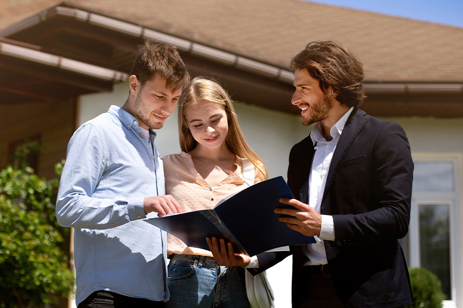 A real estate agent in a dark blazer reviews documents from a folder with a young couple standing in front of a house on a sunny day.