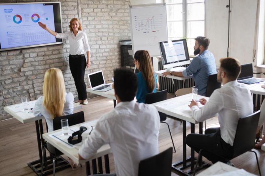 A professional instructor stands at the front of a modern, industrial-style classroom, gesturing toward a large digital display showing colorful data charts and a monthly budget. Several professionals sit at white desks with laptops, facing the instructor and taking notes in a bright, collaborative environment.