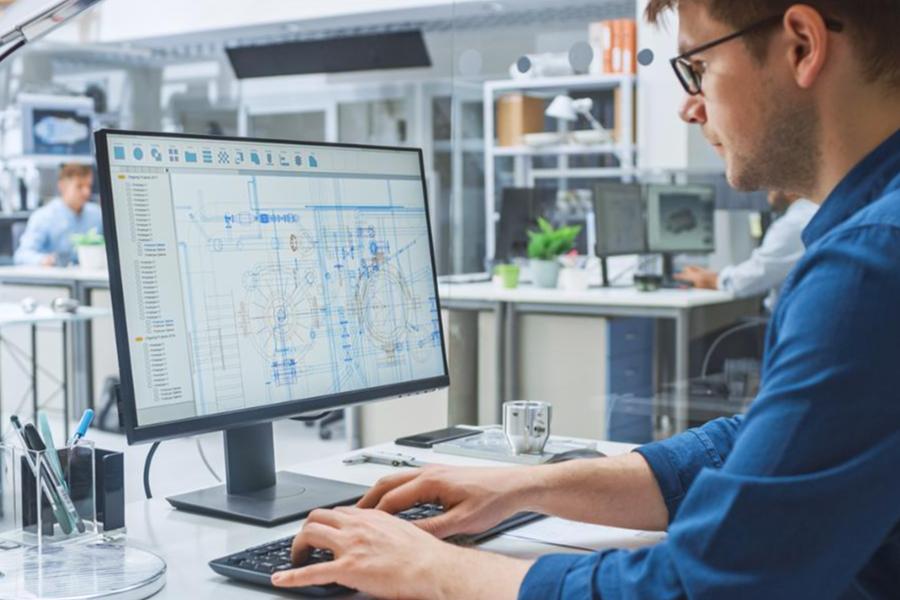 An engineer in a blue shirt using CAD software on a computer to review technical blueprints in a modern office.