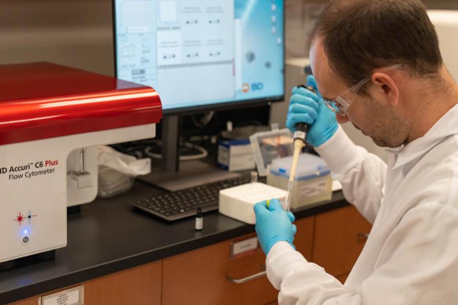 A scientist in a white lab coat and blue gloves uses a pipette next to a flow cytometer and computer monitor in a research laboratory.