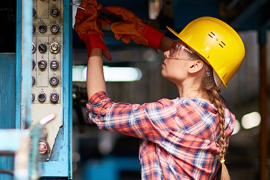 A woman in a yellow hard hat, safety glasses, and work gloves uses a screwdriver on an electrical panel in an industrial setting.