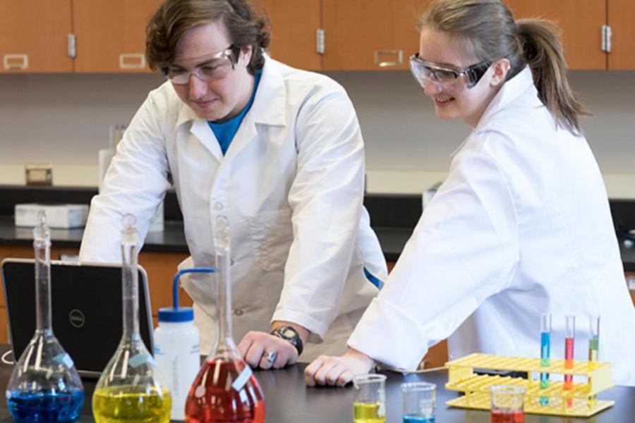 Two science students in white lab coats and safety glasses work together in a chemistry lab, reviewing data on a laptop surrounded by colorful beakers and test tubes.