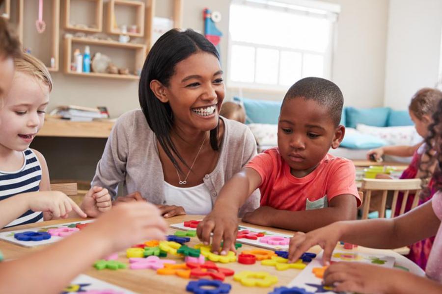 A smiling female teacher helping preschool children solve puzzles at a table in a bright classroom.