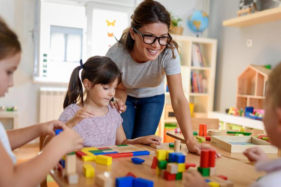 A smiling teacher in glasses observing preschool children playing with colorful wooden building blocks in a classroom.