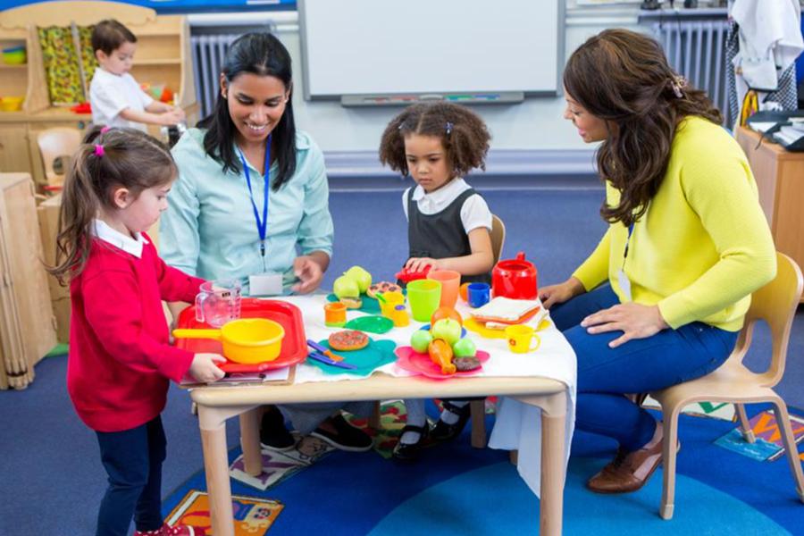 Two preschool teachers supervising a group of young children playing with toy food in a classroom.