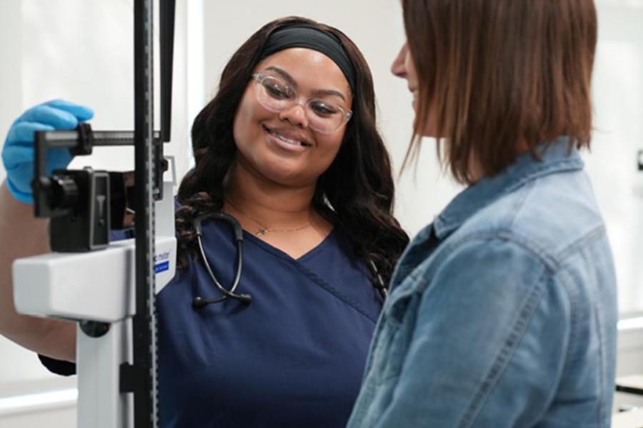 FCC medical assistant student in navy scrubs and stethoscope smiling while assisting a patient at a medical scale.