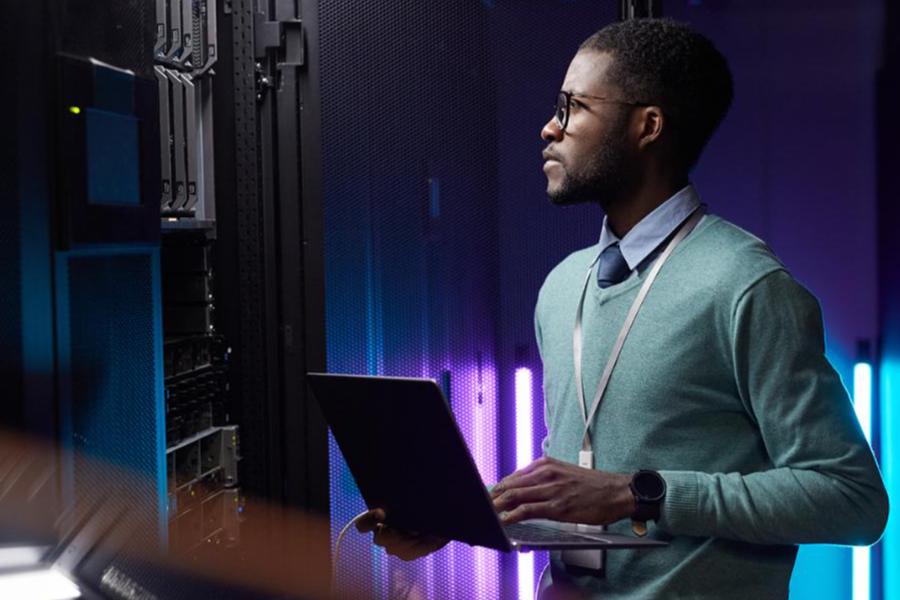A man in a green sweater and glasses looks at server racks in a dimly lit data center while holding a laptop.