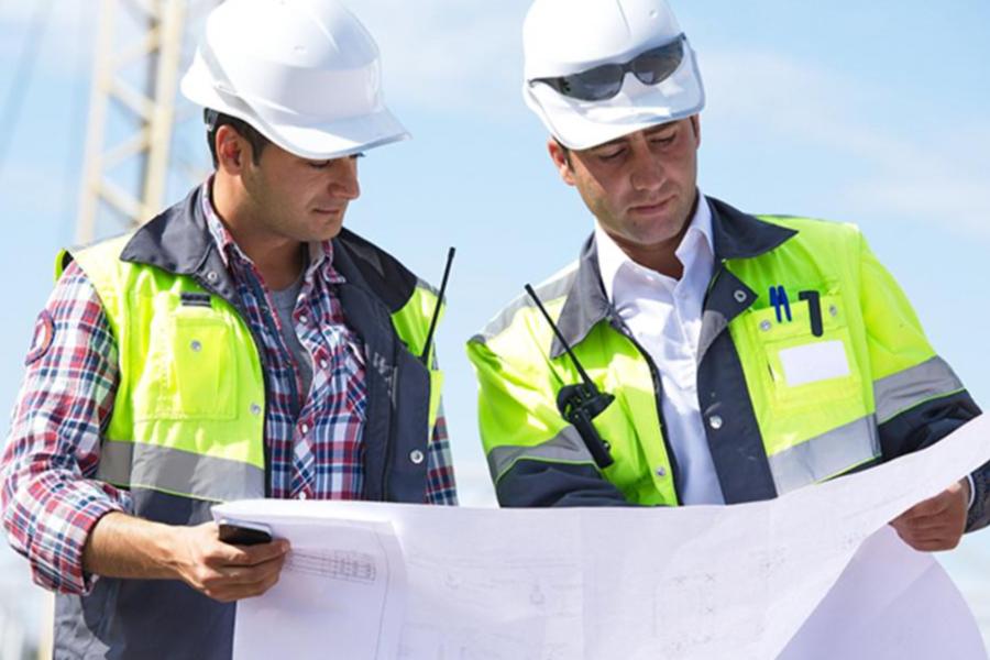 Two construction professionals in white hard hats and high-visibility safety vests review a set of large blueprints outdoors.