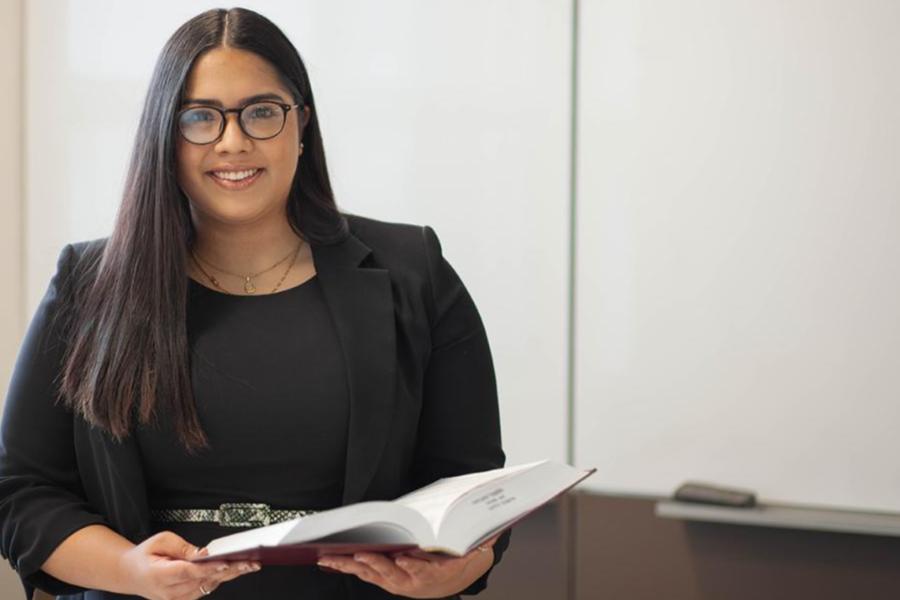 A professional portrait of a woman with glasses and long dark hair, wearing a black blazer and holding an open book in front of a whiteboard.