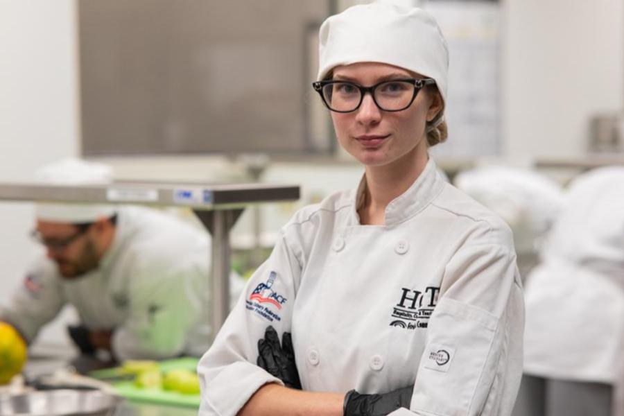 A focused female culinary student in a white chef's uniform and toque standing in a professional training kitchen.