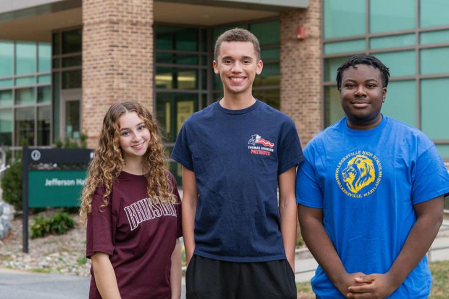 Three high school students standing outside Jefferson Hall. The young woman on the left wears a maroon t-shirt, the young man in the center wears a navy Thomas Johnson Patriots t-shirt, and the young man on the right wears a bright blue Walkersville High School t-shirt. They are smiling in front of a modern brick building and a green directional sign.