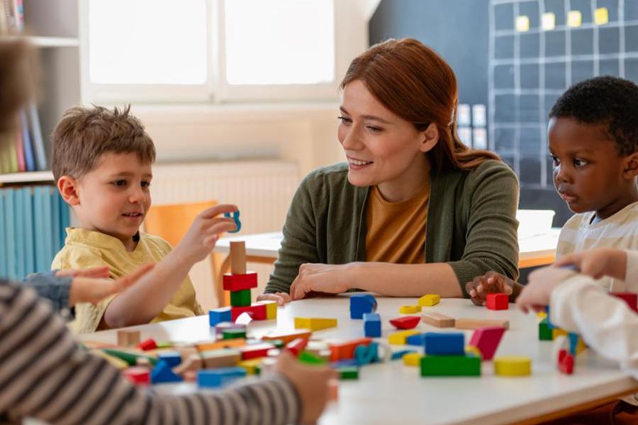 A smiling female teacher sitting at a table and supervising young children as they play with colorful wooden blocks in a classroom.
