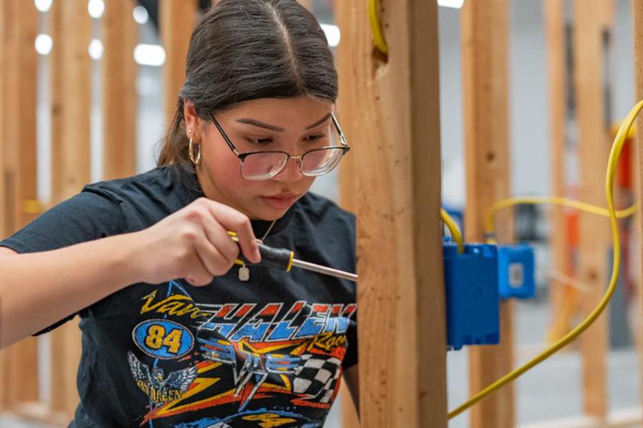 A female student wearing glasses focuses intently while using a screwdriver to work on electrical wiring in a wood-framed construction setting.