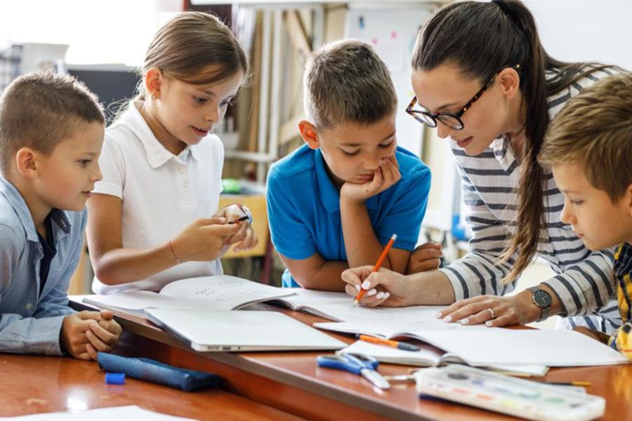 A teacher wearing glasses and a striped shirt helps four young students with their schoolwork as they gather around a desk in a classroom.