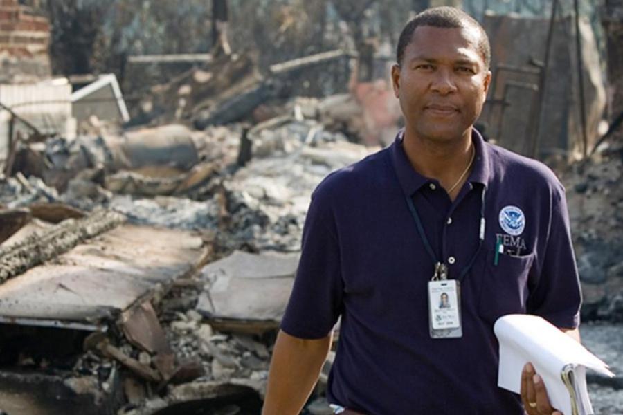 FEMA official standing in front of fire-damaged ruins holding documents.