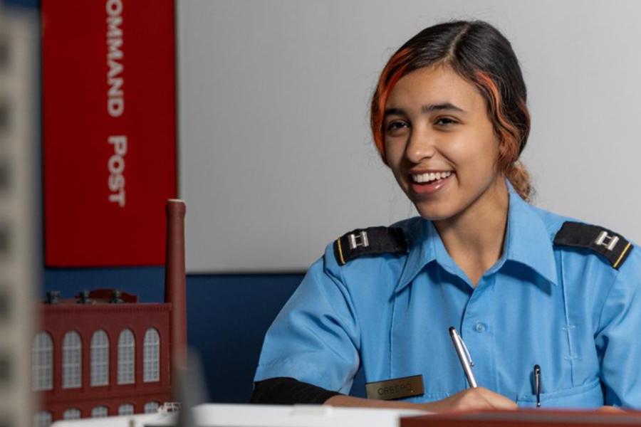 A smiling young woman in a light blue public service uniform with shoulder epaulets sits at a desk, holding a pen as if taking notes. In the background, a red sign reads COMMAND POST next to a scale model of a brick building, suggesting an emergency management training environment.