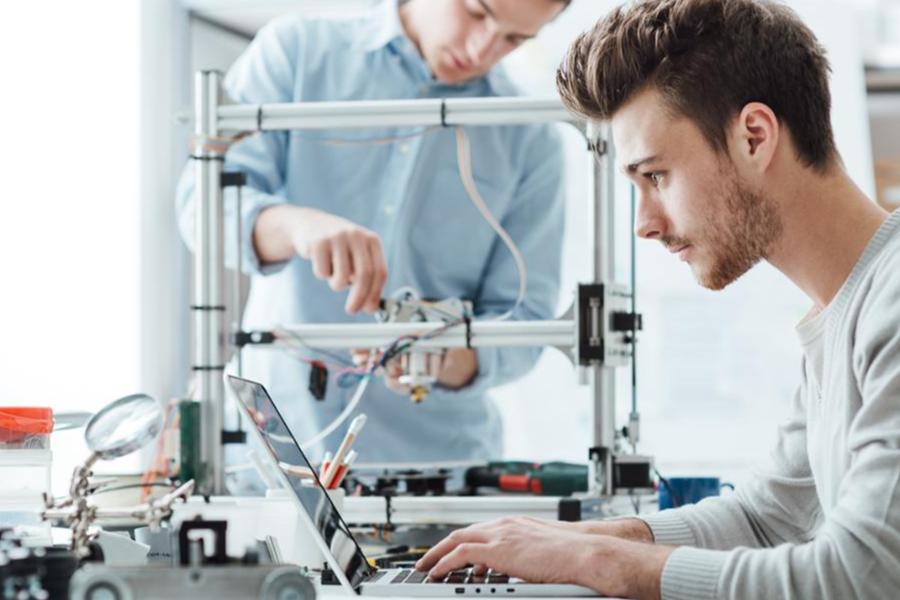 An engineering student uses a laptop to program a complex robotic assembly while a peer adjusts the mechanical framework in a bright, modern lab.