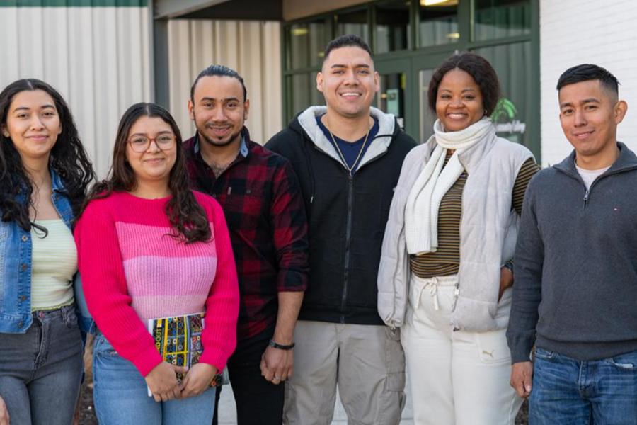 A diverse group of six smiling adult students standing together outside a Frederick Community College building. They are dressed in casual attire, including sweaters and jackets, and one student is holding a colorful notebook.