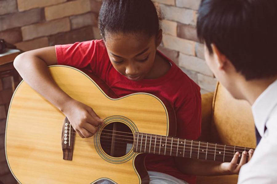 A young girl in a red shirt focuses intently as she practices playing an acoustic guitar during a lesson. An instructor is partially visible in the foreground, observing and guiding her technique in a warm, indoor setting.