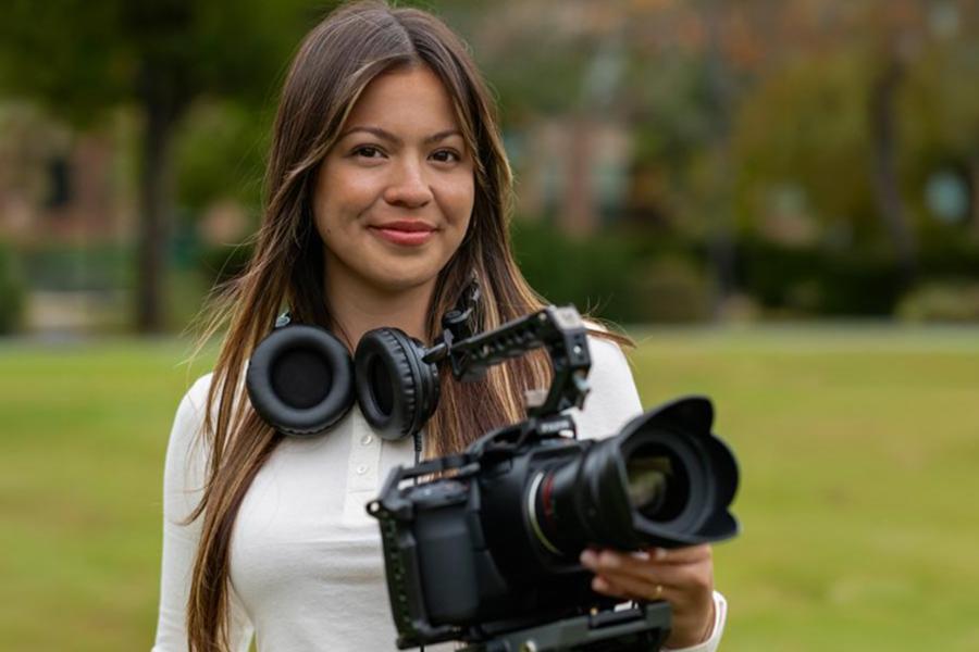 A young woman with long hair and headphones around her neck holds a professional video camera while standing in a grassy park.