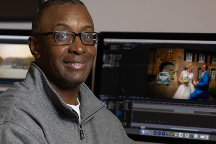 A portrait of a man with glasses smiling in front of a computer monitor showing video editing software and a wedding scene.