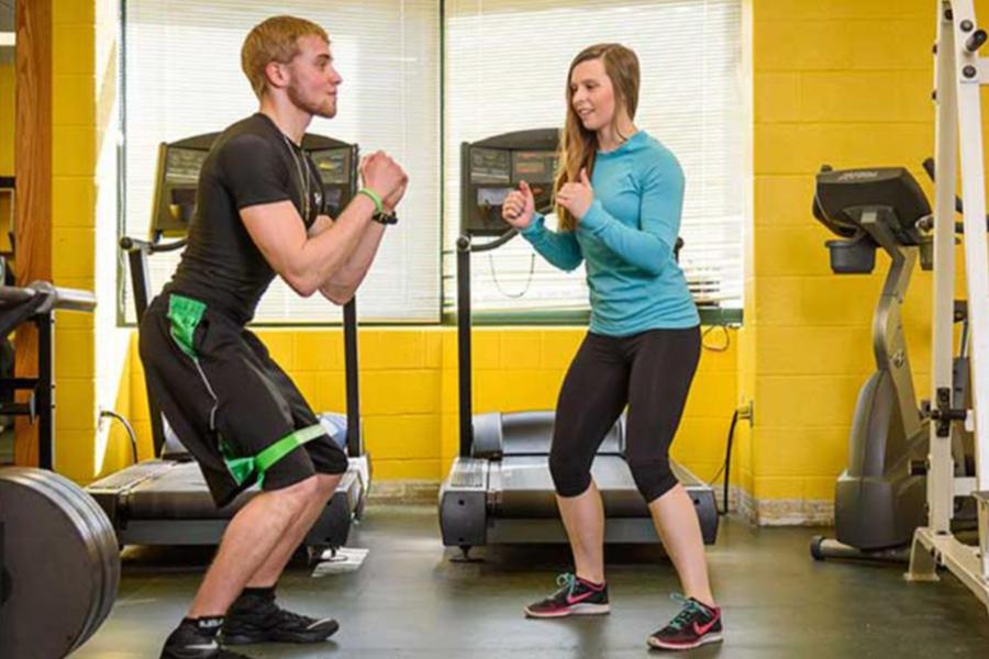 A male and female student in a bright yellow gym facility perform guided squats together in front of treadmills.