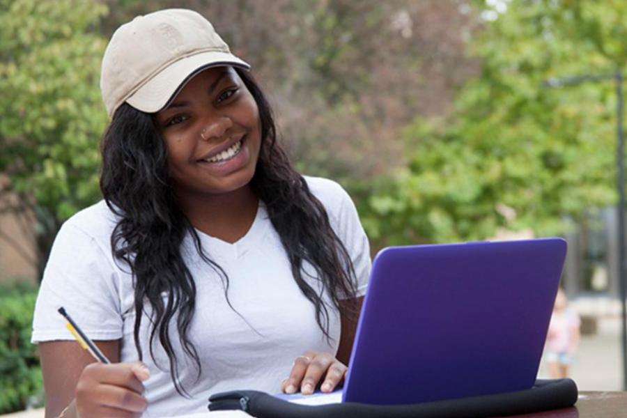 Smiling student sitting outdoors studying with a purple laptop.