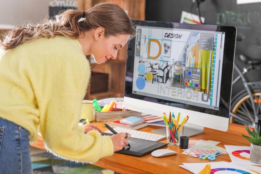 A young woman with her hair in a ponytail leans over a desk, using a stylus and drawing tablet to work on an interior design project displayed on a large iMac.