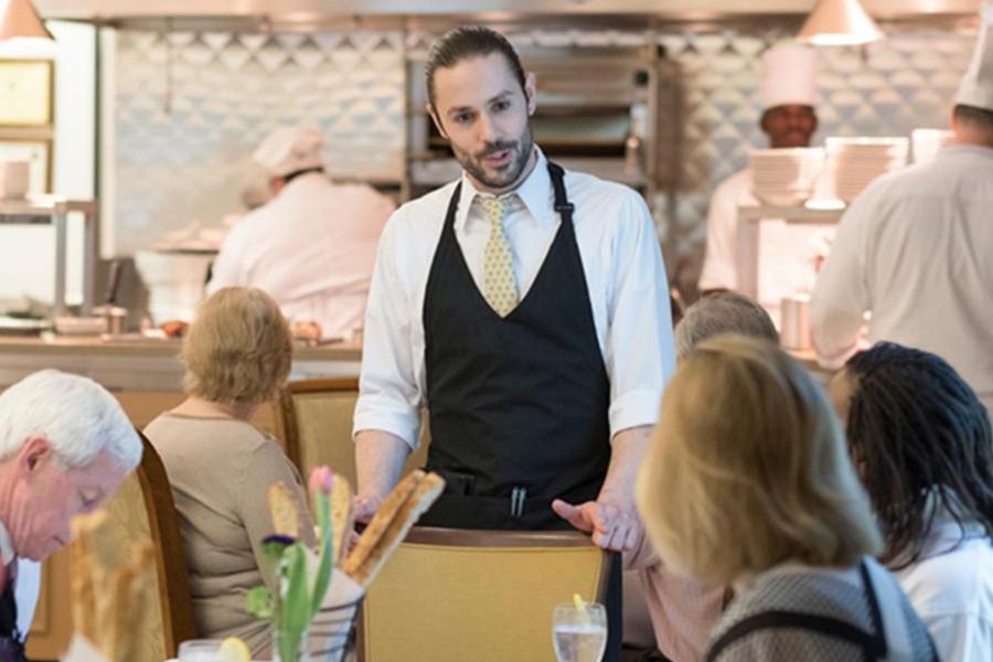 A dining room manager with a black apron and tie speaking to guests at a table in a busy restaurant with an open kitchen in the background.